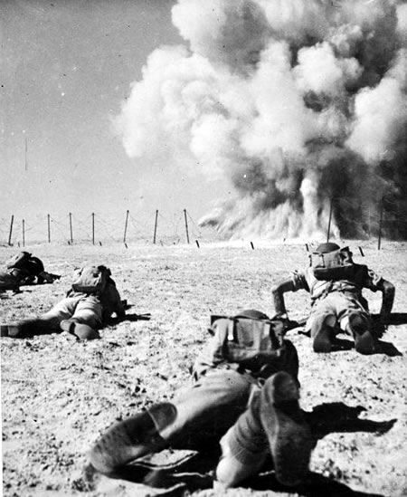 Māori Battalion troops in the North African desert keep low as a shell explodes in front of them